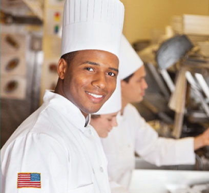 A man wearing a tall chef hat and a uniform with a backwards United States flag on the sleeve smiles at the camera.