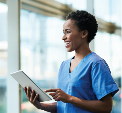 A profile shot of a nurse in blue scrubs grinning as she uses a tablet.