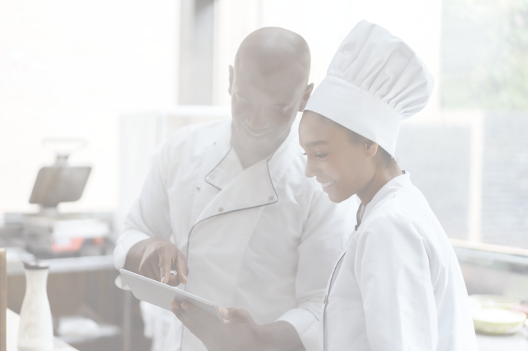 A male chef holding a tablet and a female chef wearing a puffy hat standing in a hospital kitchen automated by foodservice management software.