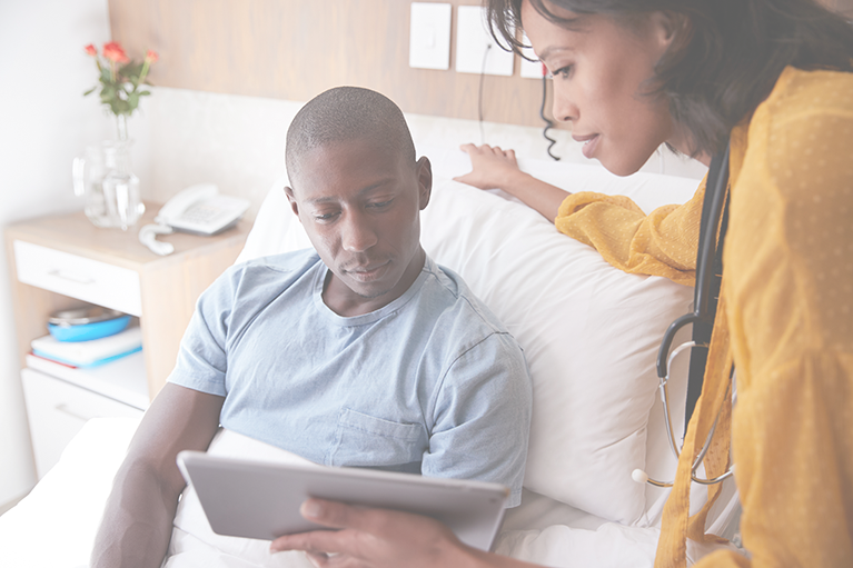 A female doctor holds a tablet as she leans over a male patient's hospital bed. They are using patient bedside ordering software to select meal options.