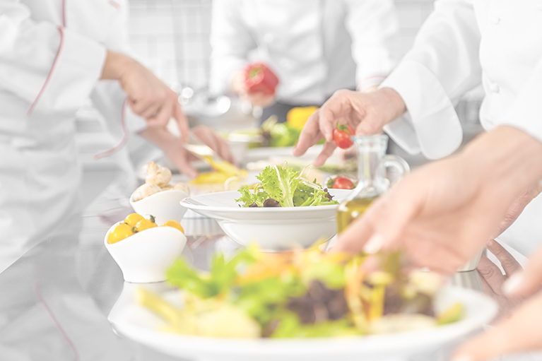 Foodservice workers assemble salads on a stainless steel worktop.