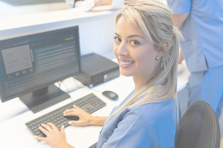 A nurse in blue scrubs sits at a computer with her hands resting on the keyboard. She is turning and smiling at the camera.