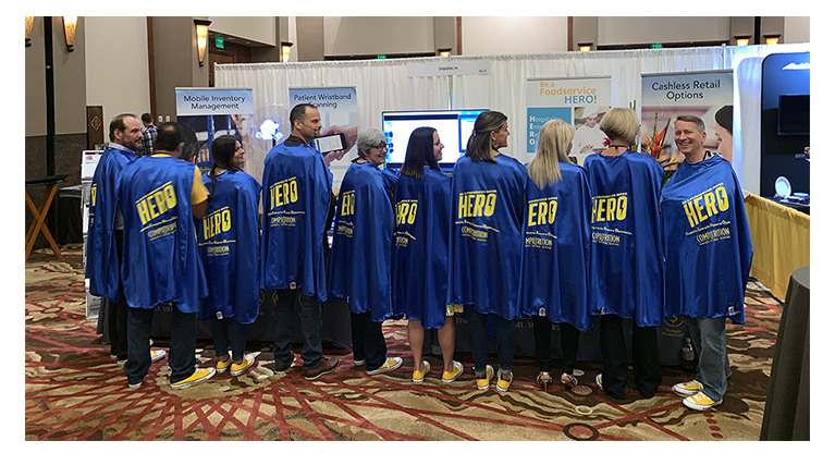 Computrition team members pose in front of a trade show booth in their H.E.R.O. capes and yellow Converse sneakers.
