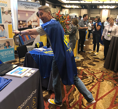 Marty Yadrick is wearing a Computrition H.E.R.O. cape and yellow Converse sneakers. He is posing in front of the CI booth at a trade show.