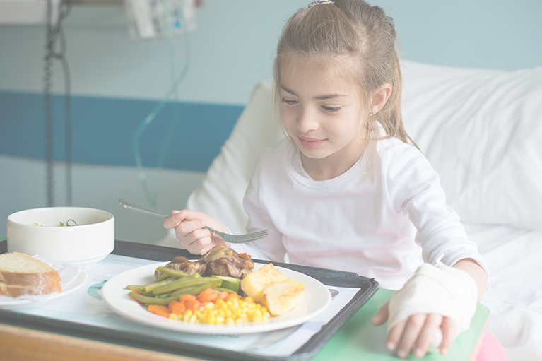 A little girl with a bandaged left hand is looking down at her hospital tray, which holds a plate of meat and veggies.